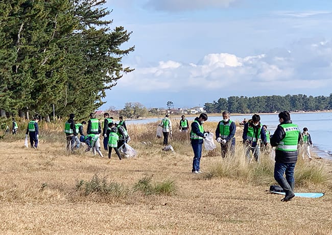 多くの参加者が緑のビブスを着用し、湖岸の草地で清掃活動を行っている様子