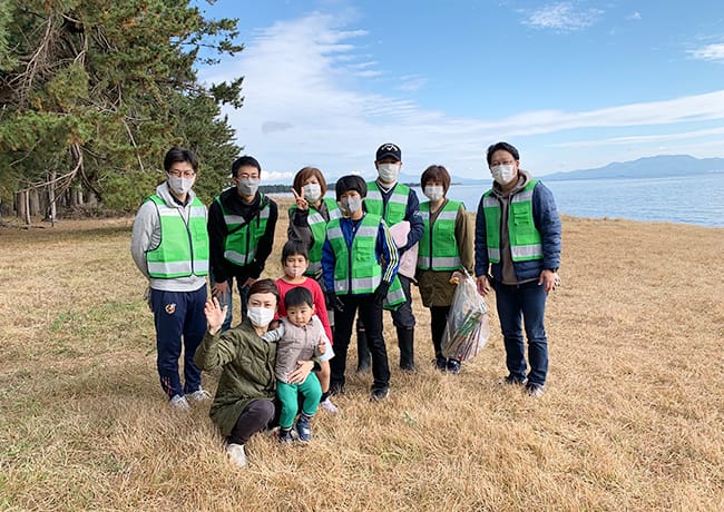 清掃活動後に、湖を背景に集合写真を撮る参加者たち。子ども連れの家族も参加している
