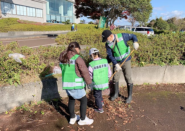 建物前で、大人と子どもが一緒に落ち葉を集め、袋に入れている清掃活動の様子