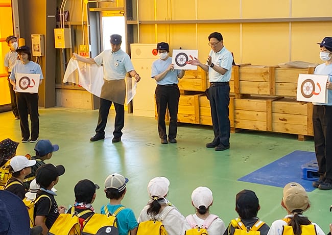 工場スタッフが児童に向けて説明を行っている様子。図を掲げながら作業内容を紹介している。