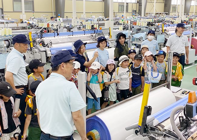 織機が並ぶ工場内で、児童たちがスタッフの説明を聞きながら見学している様子。