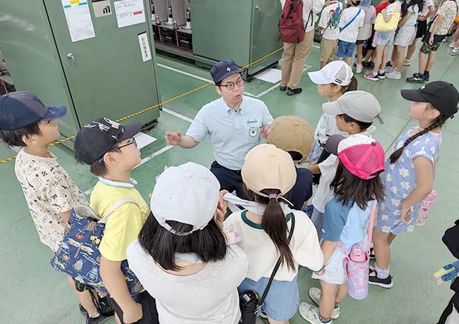 工場内でスタッフが児童たちに囲まれ、説明をしている様子。子どもたちは真剣に耳を傾けている。