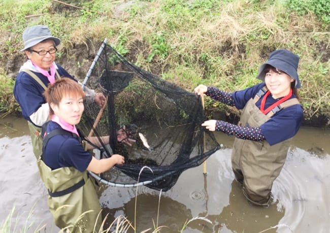 胴長を着た参加者3人が、川の中で捕まえた魚を網の中に入れて見せている様子