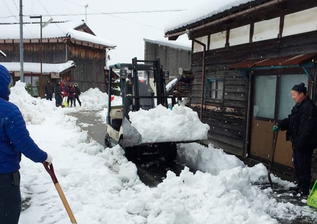 積雪の中、フォークリフトを使って雪かきをする人々の様子。周囲の住民も協力している