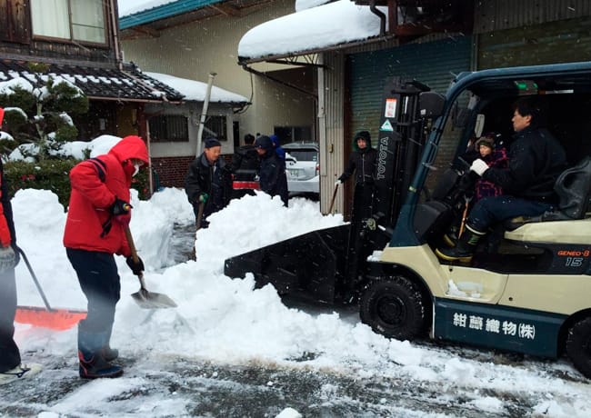 フォークリフトとスコップを使い、複数人が協力して除雪作業を行っている様子