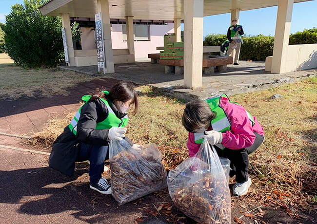 公園の東屋付近で、参加者が落ち葉を集めた袋を持ち、地面の清掃を行っている様子