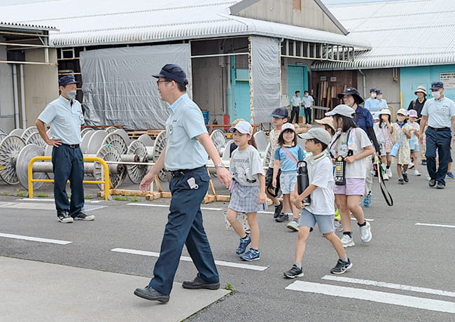 工場敷地内をスタッフに案内されながら歩く児童たちの列。屋外での見学風景。