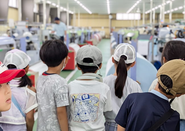 織機の並ぶ工場内で、児童たちが機械を見つめながら見学している後ろ姿。