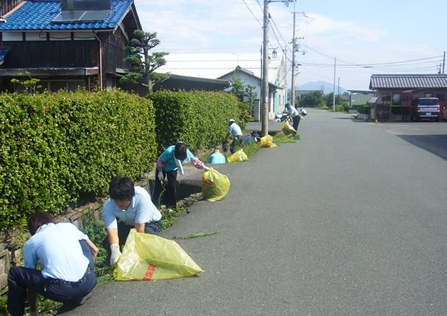 社員が道路沿いの草取りを行い、黄色いごみ袋に雑草を集めている様子